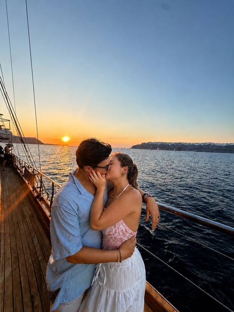 Young couple kissing on deck of a boat with orange sunset and whitewashed cliffs of Santorini in background