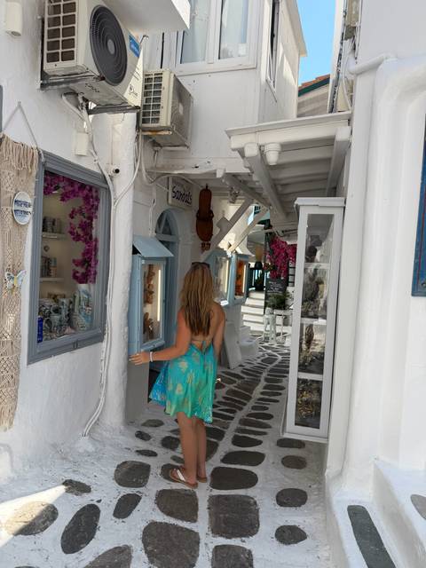 Woman entering quaint shop in flower-lined lane of Mykonos with white walls and blue shutters