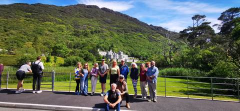 Tour group poses before the grand Kylemore Abbey set against a green mountain and blue sky.