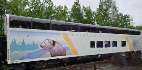 Double-deck Wilderness Express train car viewed through a rain-streaked window, featuring a painted bear and mountain scene.