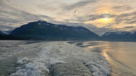 Boat wake trails toward snow-capped mountains and a golden sun breaking through dramatic clouds over icy Alaskan waters.