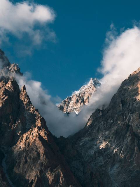 Snow-capped rocky summit piercing low swirling clouds against a blue sky.