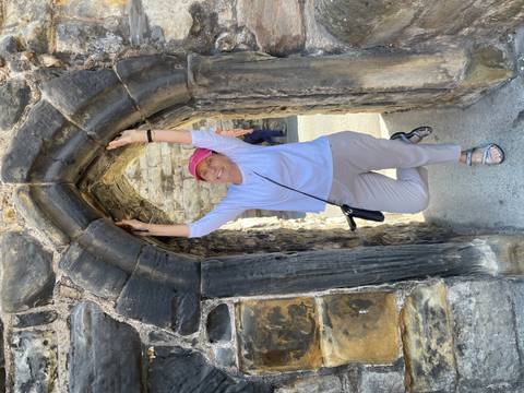 Smiling woman in pink cap playfully hangs from a narrow stone archway in an ancient ruin.