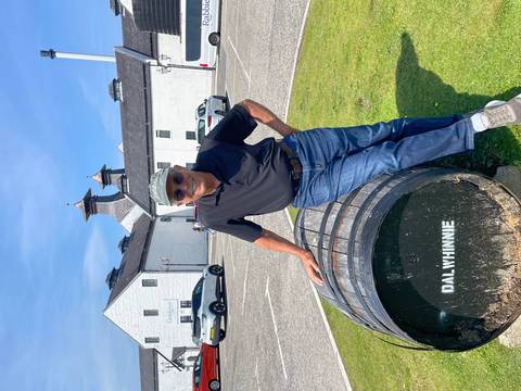 Man poses beside a whisky barrel labeled “DALWHINNIE” outside the white-washed distillery buildings.