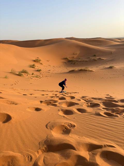 Woman clambers up an orange dune in the Sahara, footprints trailing behind in rippled sand.