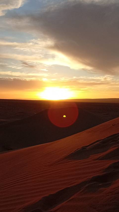 Sun sinks below a desert horizon casting golden light and lens flare over shadowed dunes.