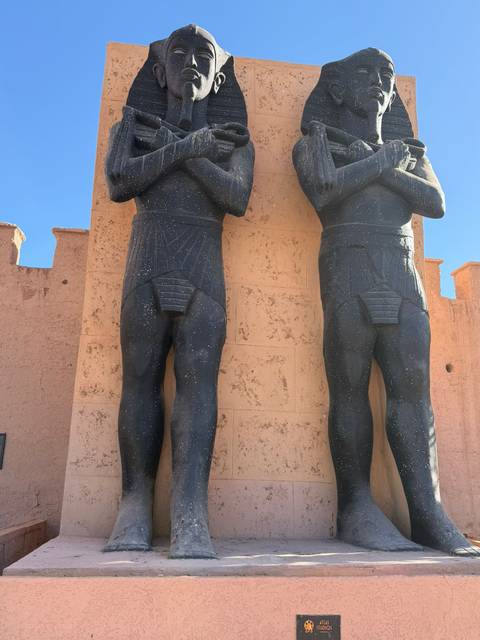 Two towering black statues of ancient Egyptian soldiers stand before a pinkish adobe wall at a film studio.