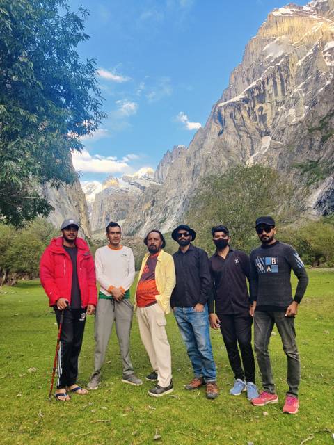 Group of six friends standing in lush alpine meadow with towering granite spires rising behind.