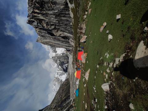 Colorful expedition tents dotting a grassy alpine campsite beneath towering snow-streaked cliffs under a dramatic sky.