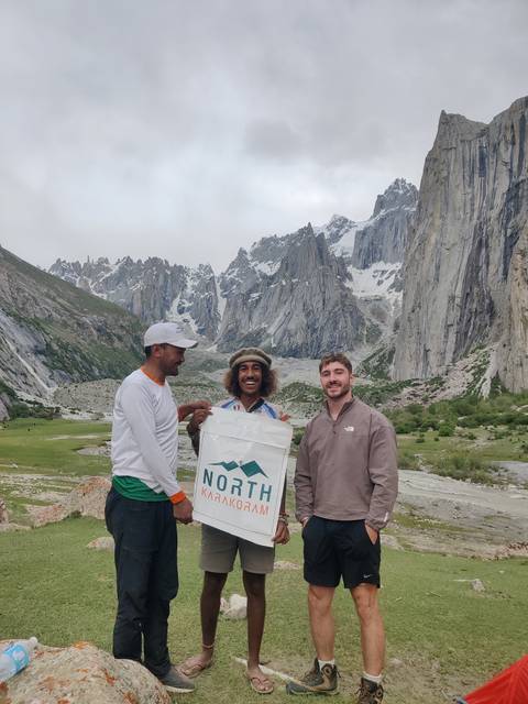 Three trekkers smiling with North Pakistan banner in front of dramatic jagged peaks and glacier valley.
