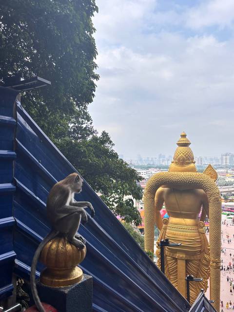Monkey sitting on Batu Caves stair rail overlooking giant golden Murugan statue and city skyline
