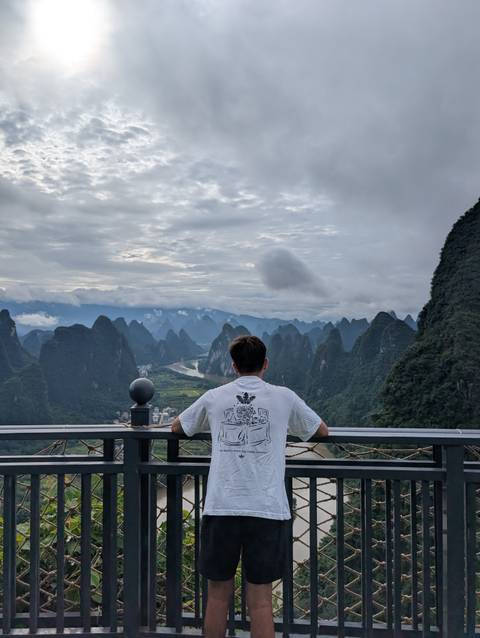 Traveller gazes out over dramatic karst mountains and winding river valleys under a cloudy sky.