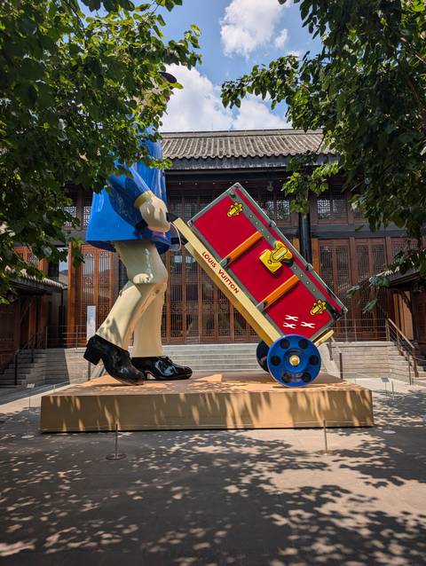 Large colourful Louis Vuitton trunk sculpture pushed by a stylised figure outside a wooden building.