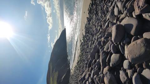 Sunlit rocky beach with smooth stones in foreground and shimmering sea under a bright sky.