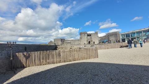 Stone walls and towers of a medieval riverside castle with a few visitors walking across a gravel courtyard on a sunny day.