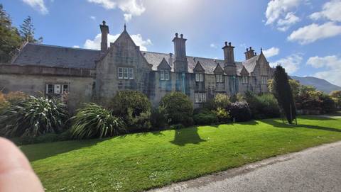 Historic manor house with tall chimneys and stone façade surrounded by manicured lawn and shrubs on a bright day.
