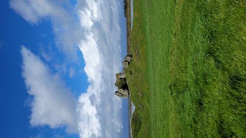 Rolling green pasture with a rocky hillock beneath a dramatic sky of sun and clouds.