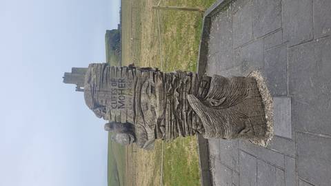 Carved wooden monument reading "Cliffs of Moher" with grassy cliffs visible in the distance.