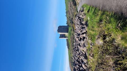 Ruined stone tower standing on a grassy rise with boulder field and winding path under a vivid blue sky.