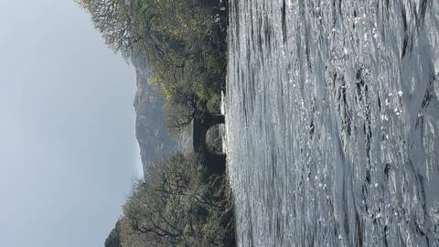 Wide river with fast-flowing water and an old stone arch bridge framed by spring trees and distant hills.