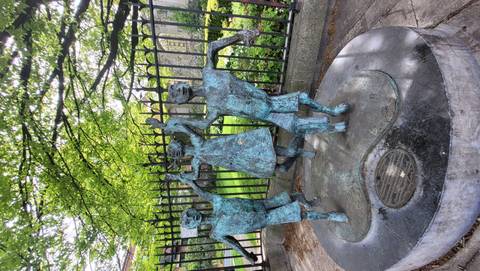 Bronze sculpture of three dancing children beneath leafy trees beside an iron fence in a city park.