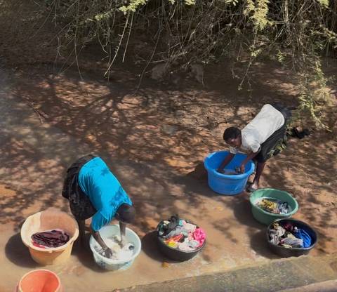 Two local women hand-washing clothes in shallow muddy river using plastic basins.