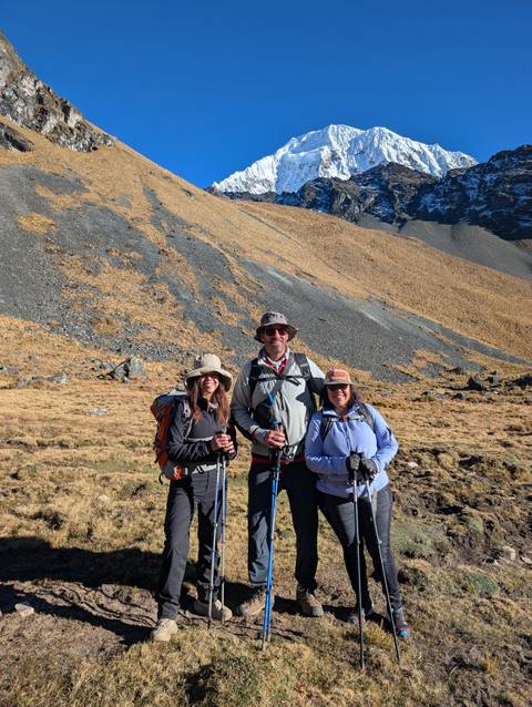 Three trekkers with poles pause on a grassy Andean slope with scree and peaks behind.