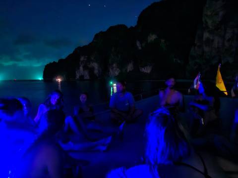 Small group of young travelers sitting on a dark boat at night, illuminated by deep blue lights against towering limestone cliffs.