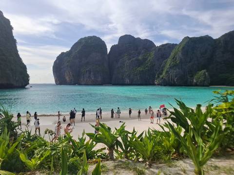 Panoramic view of a tropical beach with clear aquamarine water, lush green cliffs and visitors enjoying the shoreline.