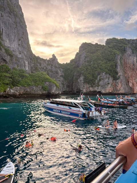 Speedboats and long-tail boats gather in a turquoise cove surrounded by steep limestone walls and jungle vegetation.