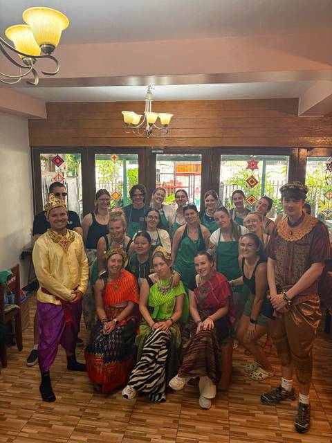 Large tour group wearing aprons and Thai cultural outfits posing inside a cooking classroom.