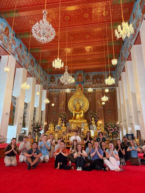 Opulent Thai temple interior featuring a large golden Buddha statue surrounded by flowers, chandeliers and intricate murals.