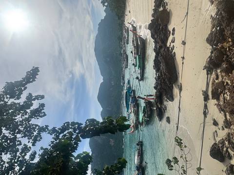Long-tail boats anchored in turquoise water off a small sandy beach with jungle-covered mountains in the background.