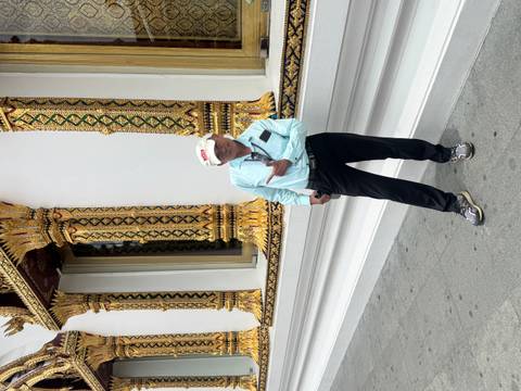 A smiling tourist poses in front of an intricately decorated Thai temple facade featuring gold and green motifs.