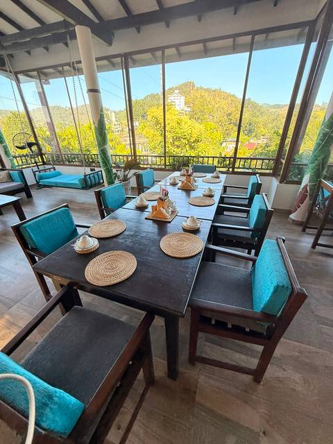 Open-air dining area with a long dark wooden table, turquoise cushions and views of greenery outside.