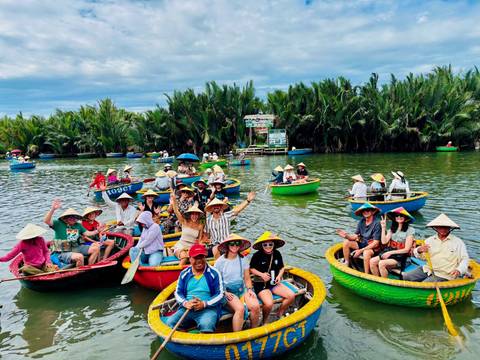 Dozens of travelers wearing conical hats sit in round basket boats on a calm tropical river lined with dense palms.
