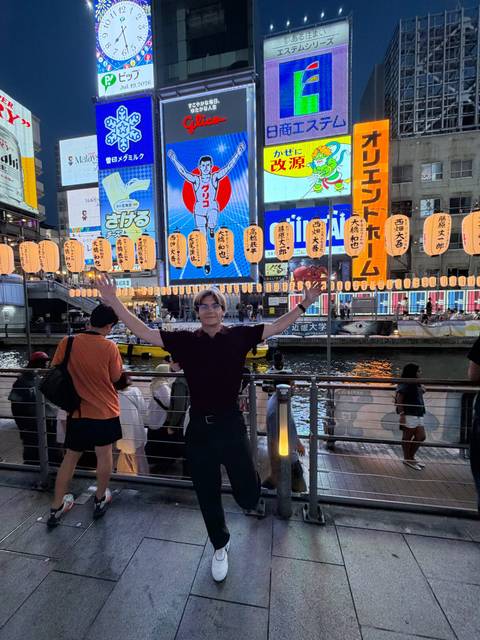 Nighttime scene at Osaka’s vibrant Dotonbori canal with a smiling tourist posing under rows of glowing lanterns.
