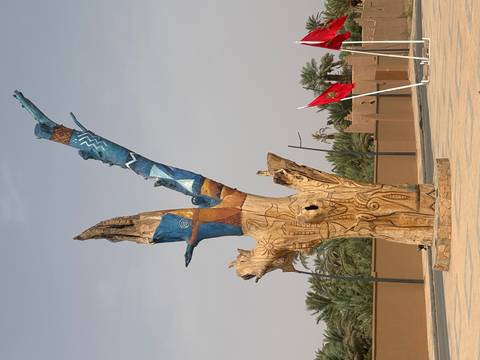 Carved and painted dead tree sculpture standing in a desert town square beside a Moroccan flag.