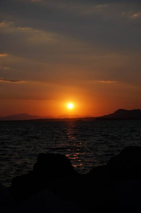 Brilliant orange sunset casting warm glow over calm sea with silhouetted mountains on the horizon.