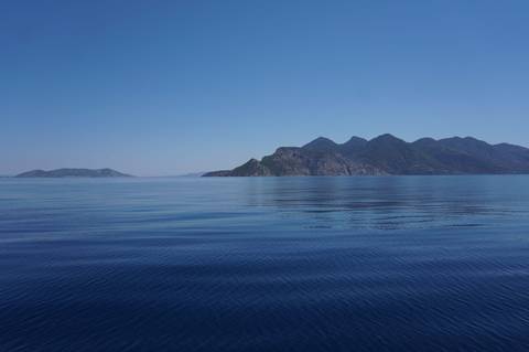 Glass-calm blue sea reflecting distant rugged islands under a clear midday sky.