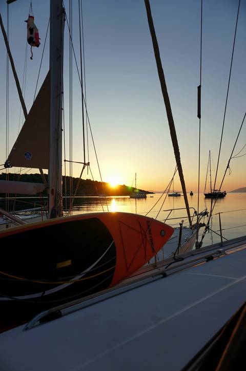 Sailboat deck at sunrise with other anchored yachts and golden light reflecting on tranquil water.