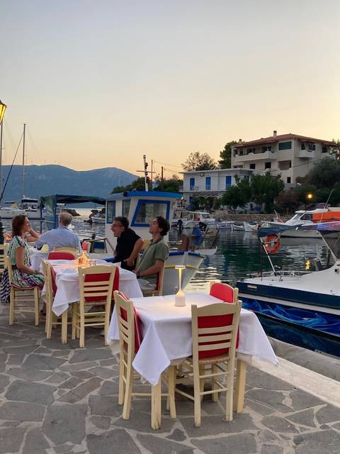 Outdoor waterfront restaurant at dusk with diners seated beside small fishing boats in a tranquil harbor.