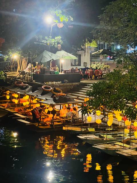 Night view of lantern-lit boats and riverside dining decks along a canal with people enjoying the evening.