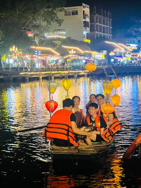 Tourists wearing life vests sit in a lantern-decorated boat on a shimmering river at night, city lights behind.