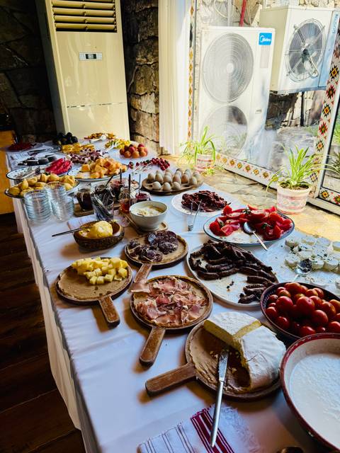 A long buffet table is covered with a rustic spread of local cheeses, meats, fruits and breads, lit by daylight from a window.