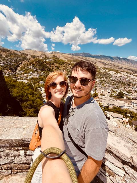 A couple takes a selfie atop a fortress, overlooking a sprawling historic town and surrounding mountains under bright sun.