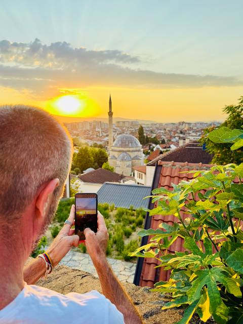 At sunset a traveler photographs a domed mosque and cityscape glowing under an orange sky from a leafy terrace.