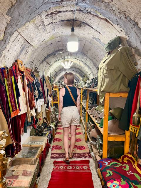 Inside a bunker-style museum corridor, a woman examines vintage military uniforms and artifacts displayed on wooden shelves.