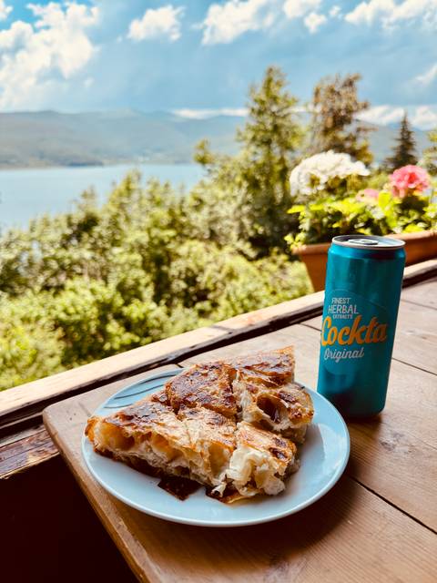 Plate of flaky pastry and a blue can of Cockta herbal soda on a wooden balcony rail overlooking green trees and a calm lake
