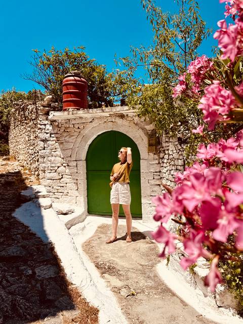 Young woman posing in sunlight before a rustic stone arch with a bright green wooden door, pink flowers framing the scene
